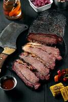 Fresh Brisket BBQ beef sliced for serving against a dark background with sauce, hot peppers and corn. photo