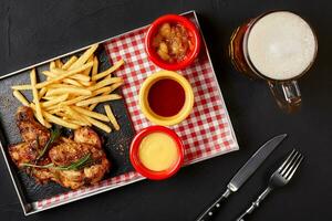 horneado pollo alas con francés papas fritas en un hierro bandeja con vaso de cerveza en negro antecedentes foto