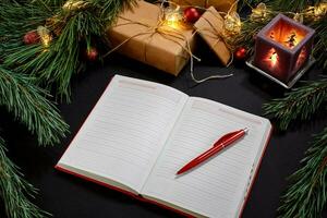 Red Christmas balls and notebook lying near green spruce branch on black background top view. Space for text photo