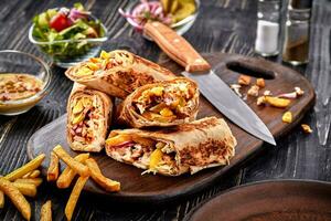 Shawarma chicken roll in a pita with fresh vegetables, cream sauce and french fries on wooden background. Selective focus photo