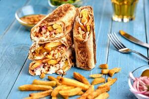 Shawarma chicken roll in a pita with fresh vegetables, cream sauce and french fries on wooden background. Selective focus photo