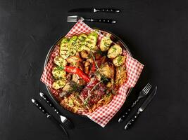 Assorted delicious grilled meat with vegetables and bbq sauce on an iron tray on black background close up. Big set of Hot Meat Dishes. Top view photo