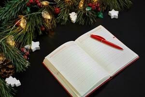 Christmas balls and notebook lying near green spruce branch on black background top view. Space for text photo