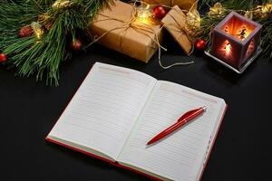 Red Christmas balls and notebook lying near green spruce branch on black background top view. Space for text photo