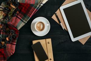 Above view of Smart phone, tablet, scarf in a cage, glasses with notebook and cup of latte coffee on black wooden background. photo
