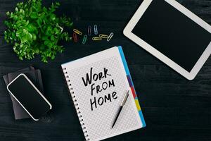 Top view of open notebook written with WORK FROM HOME inscription. Green flower, tablet, colored paper clips, smart, pen and notebook on wooden background. photo