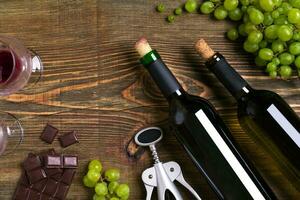 Red and white wine bottles, grape, chocolate and glasses over wooden table. Top view with copy space photo