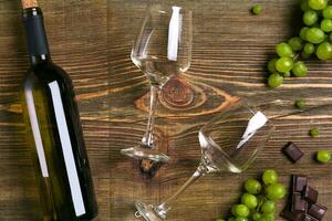 White wine bottle, grape, chocolate and glasses over wooden table. Top view with copy space photo