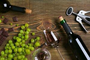 Red and white wine bottles, grape, chocolate and glasses over wooden table. Top view with copy space photo