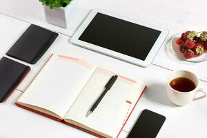 Office table desk with set of supplies, white blank notepad, cup, pen, tablet, glasses, flower on white background. Top view photo