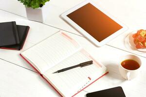 Office table desk with set of supplies, white blank notepad, cup, pen, tablet, flower on white background. Top view photo