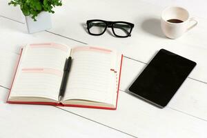 Office table desk with set of supplies, white blank notepad, cup, pen, tablet, glasses, flower on white background. Top view photo