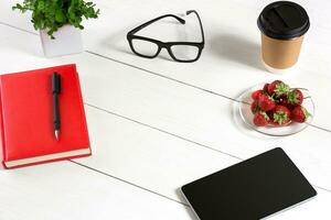 Office table desk with set of supplies, red notepad, cup, pen, tablet, glasses, flower on white background. Top view photo