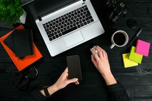 Female hands holding a smart and typing on the keyboard of a laptop on black wooden table. photo