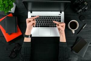 mujer trabajando a el oficina mesa. parte superior ver de humano manos, ordenador portátil teclado, un taza de café, teléfono inteligente, cuaderno y un flor en un de madera mesa antecedentes foto