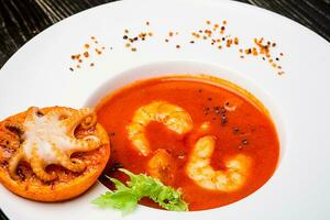 Bowl of tomato soup with shrimps with a small octopus baked on an orange slice on a black wooden background, top view photo