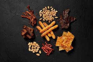 Top view of various beer snacks like peanut, crackers, sausages, chips, jerky, cheese sticks on dark desk. photo