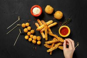 Mushrooms in batter, cheese balls, cheese sticks and cheese rings in batter, on black background, with herbs, sauce, ketchup and pepper. Top view photo