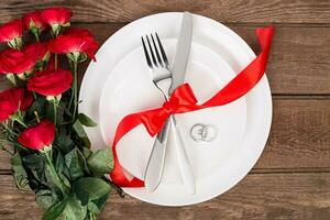 San Valentín día cena mesa ajuste con rojo cinta, rosas, cuchillo y tenedor anillo terminado roble antecedentes. foto