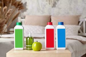 Three colorful plastic containers with detergents, pump bottle with soap and green apple on wooden bedside table in bedroom. Close up, copy space photo