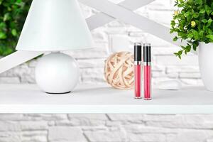 Two pink lip glosses on white rack next to a lamp, green plant in pot and round element of decor against brick wall. Close up, copy space photo