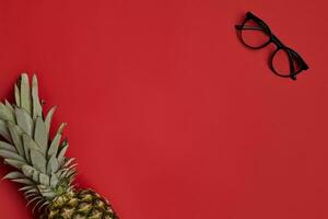 Stylish glasses with black frames and transparent lenses and ripe pineapple against red studio background. Close up, copy space. Top view photo