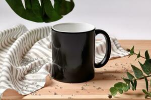 Black porcelain mug on wooden desktop next to striped tablecloth, scattered crystals and green twigs against white background. Close up, copy space photo