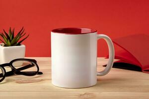 White porcelain mug on wooden desktop next to notepad, glasses and green succulent in pot against red studio background. Close up, copy space photo