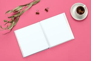 Empty open notebook, two golden binder clips, cup of coffee and green twig on pink background. Stationery, supplies for office. Close up, copy space photo