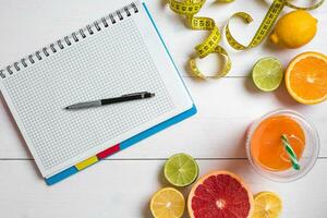 Fresh juice in glass from citrus fruits - lemon, grapefruit, orange, notebook with pencil on white wooden background photo