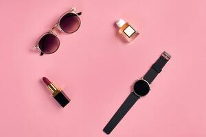 rojo lápiz labial, perfume botella con blanco etiqueta, reloj con negro marcar y elegante Gafas de sol en contra rosado antecedentes. cerca arriba, Copiar espacio foto