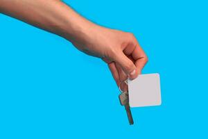 Hand of unknown male is holding a key with blank white square plastic key fob on metal ring against blue studio background. Close up, copy space photo