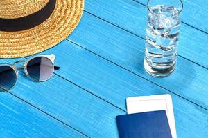 Straw hat, sunglasses, glass of water with ice cubes and passport with ticket on blue wooden background. Travelling and tourism. Close up, copy space photo