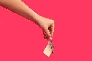 Hand of unknown woman is holding a key with blank white square plastic key fob on metal ring against pink studio background. Close up, copy space photo