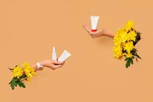 Two hands with yellow chrysanthemums holding white tubes of cream or gel and small pump bottle without logo. Blank branding area. Copy space photo