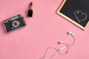 Camera, red lipstick, white earphones and small chalkboard with heart drawn on it against pink background. Close up, copy space, top view photo