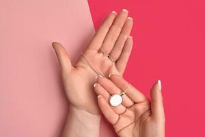 Hands of unknown young lady are holding shiny silver or platinum blank pendant on a chain against colorful studio background. Close up, copy space photo