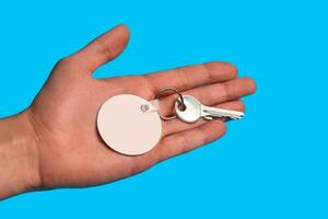 Key with blank white plastic keychain on metal ring on palm of unknown man posing against blue studio background. Close up, copy space photo