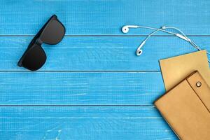 Black sunglasses, white earphones, brown craft paper sheet and leather folder on blue wooden desktop or background. Close up, copy space photo