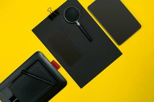 Flat lay display of business office gadgets with notepad, cup, pen, develop, glasses and etc. Top view. Black office objects on a yellow background photo