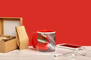 Ceramic colorful cup, cardboard box with small paper bags and smartphone with earphones on white desktop against red background. Close up, copy space photo