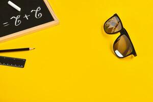 Black sunglasses, pencil, ruler and example two plus two written by chalk on a small chalkboard, yellow background. Close up, copy space photo