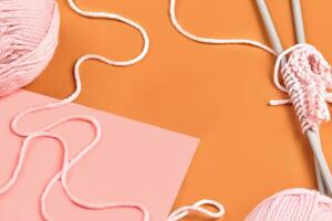 Two pink skeins of yarn and knitting on long needles, empty sheet of paper on orange background. Close up, copy space photo