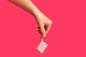 Hand of unrecognizable female is holding a key with empty white square plastic key fob on metal ring against pink background. Close up, copy space photo