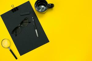 Flat lay display of business office gadgets with notepad, cup, pen, develop, glasses and etc. Top view. Black office objects on a yellow background photo