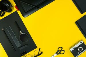 Flat lay display of business office gadgets with notepad, vintage camera, pen, develop, glasses and etc. Top view. Black office objects on a yellow background photo