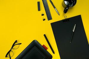 Flat lay display of business office gadgets with notepad, cup, pen, develop, glasses and etc. Top view. Black office objects on a yellow background photo