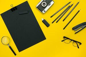 Flat lay display of business office gadgets with notepad, vintage camera, pen, develop, glasses and etc. Top view. Black office objects on a yellow background photo