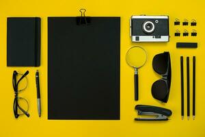 Flat lay display of business office gadgets with notepad, vintage camera, pen, develop, glasses and etc. Top view. Black office objects on a yellow background photo