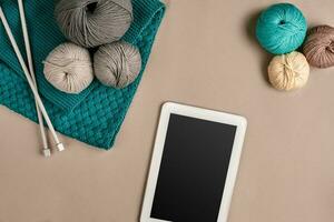 Grey and turquoise knitting wool, knitting needles and a tablet with a black screen on beige background. Top view. Copy space photo